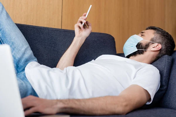 Concentration sélective de l'homme dans le masque médical à l'aide d'un smartphone près d'un ordinateur portable sur le canapé — Photo de stock