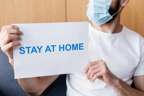 Cropped view of man in medical mask holding card with stay at home lettering on couch — Stock Photo