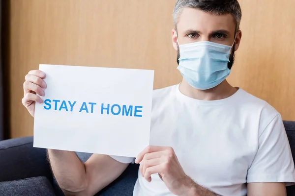 Hombre con máscara médica que sostiene la tarjeta con la estancia en casa letras y mirando a la cámara en el sofá - foto de stock