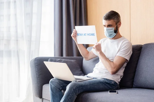 Teleworker in medical mask holding card with stay at home lettering near laptop and smartphone on couch — Stock Photo