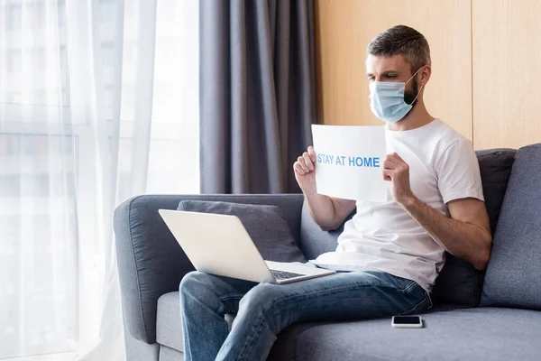 Homme en masque médical tenant la carte avec rester à la maison lettrage tout en regardant ordinateur portable sur le canapé — Photo de stock