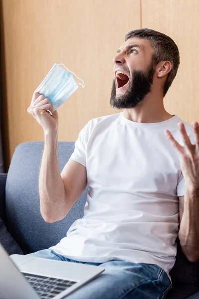 Selective focus of angry man holding medical mask near laptop on couch — Stock Photo