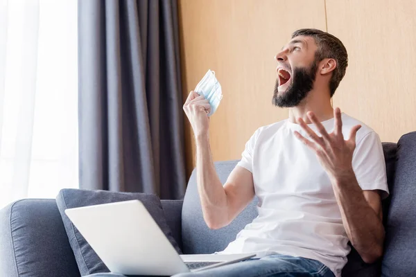 Uomo arrabbiato urla mentre tiene la maschera medica vicino al computer portatile sul divano — Foto stock