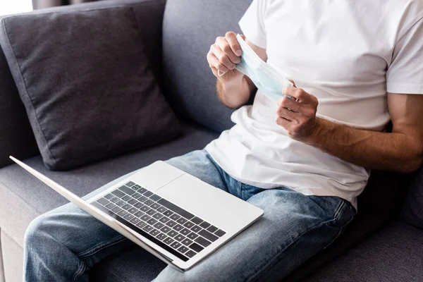 Cropped view of freelancer holding medical mask near laptop on couch — Stock Photo