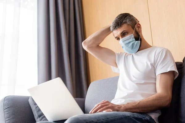 Freelancer en máscara médica usando laptop en sala de estar - foto de stock