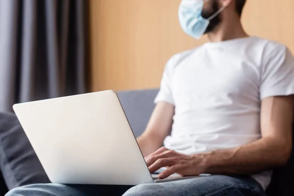 Selective focus of freelancer in medical mask using laptop on sofa at home — Stock Photo