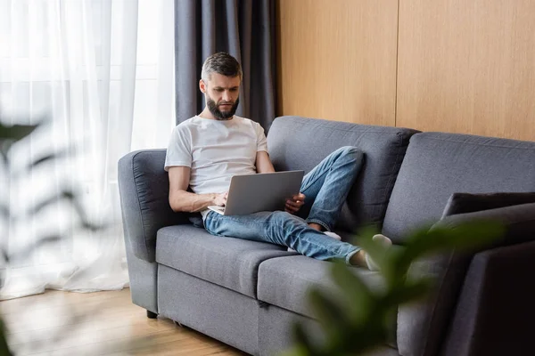 Selektiver Fokus eines hübschen Telearbeiters mit Laptop, während er zu Hause auf der Couch sitzt — Stockfoto