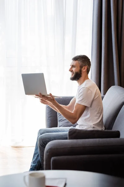 Vista lateral de sonriente freelancer tener video chat en el ordenador portátil en la sala de estar - foto de stock