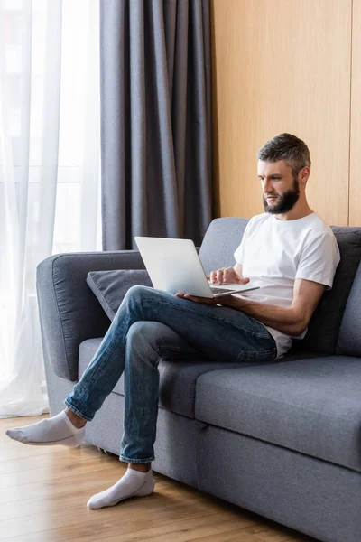 Freelancer usando laptop mientras está sentado en el sofá en casa - foto de stock