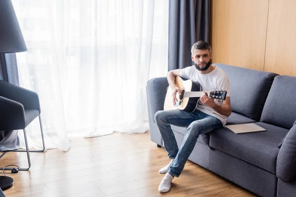 Bell'uomo che suona la chitarra acustica vicino al computer portatile sul divano a casa — Foto stock