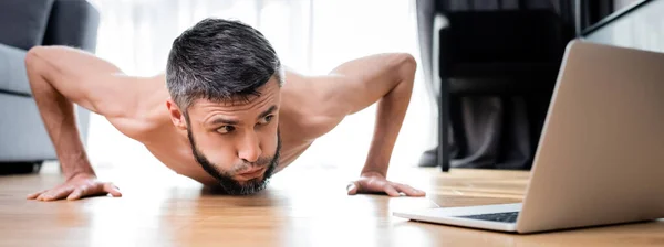 Cosecha panorámica del hombre sin camisa haciendo flexiones cerca de la computadora portátil en el suelo - foto de stock