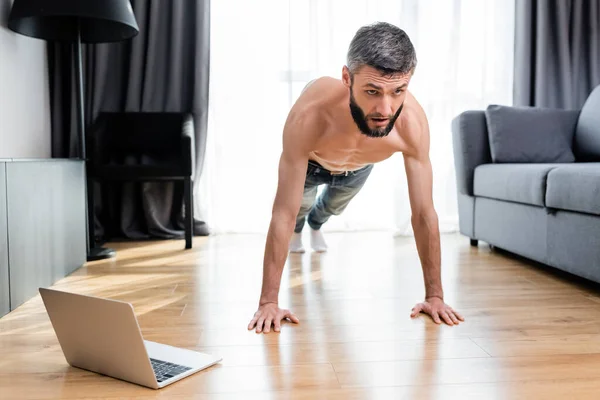 Musculoso hombre haciendo flexiones en el suelo cerca de la computadora portátil en la sala de estar - foto de stock