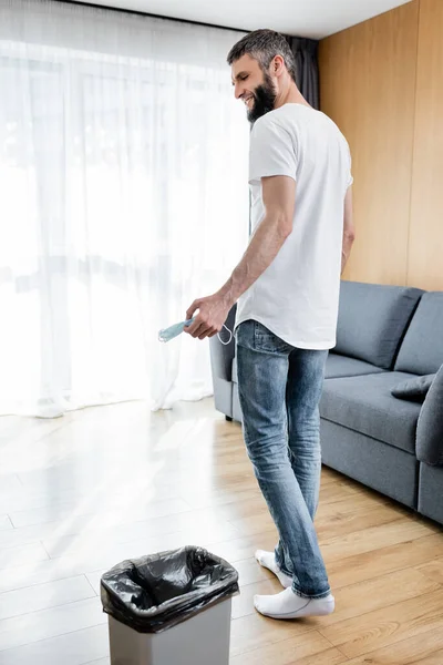 Vista trasera del hombre sonriente sosteniendo la máscara médica cerca del bote de basura en la sala de estar - foto de stock