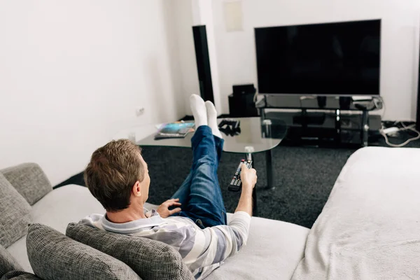 Homem de jeans segurando controle remoto perto de tela de tv em branco em casa — Fotografia de Stock