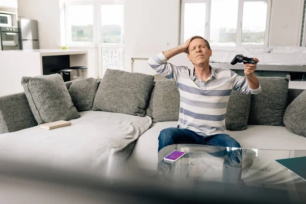 KYIV, UKRAINE - APRIL 14, 2020: selective focus of upset man touching head and holding joystick near smartphone with instagram app on coffee table — Stock Photo