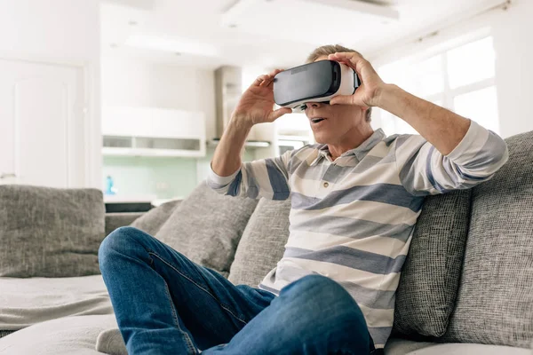 Homme surpris toucher casque de réalité virtuelle tout en étant assis sur le canapé à la maison — Photo de stock