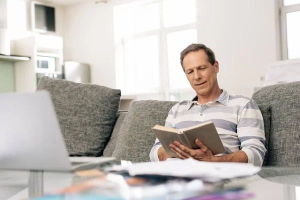 Selective focus of handsome man smiling while reading book in living room — Stock Photo