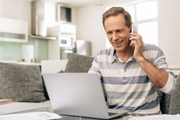 Happy freelancer talking on smartphone and looking at laptop at home — Stock Photo