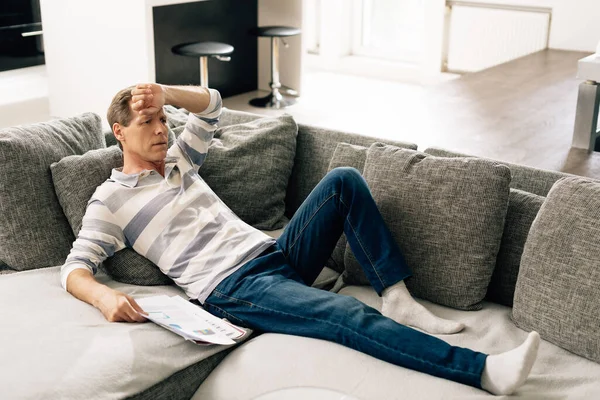 Hombre cansado sosteniendo gráficos y gráficos mientras está acostado en el sofá y tocando la cabeza - foto de stock