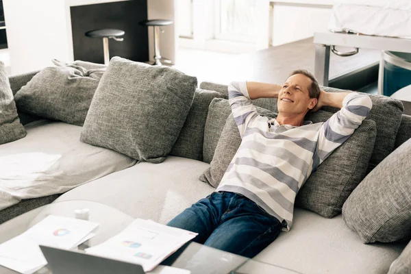 Fröhlicher Mann entspannt sich auf Sofa in der Nähe von Diagrammen und Grafiken auf Couchtisch — Stockfoto