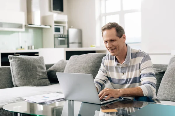 Hombre alegre sonriendo mientras usa el ordenador portátil cerca de la tarjeta de crédito en la sala de estar - foto de stock