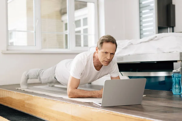 Hombre haciendo ejercicio en la alfombra de fitness mientras ve en línea de entrenamiento en el ordenador portátil - foto de stock