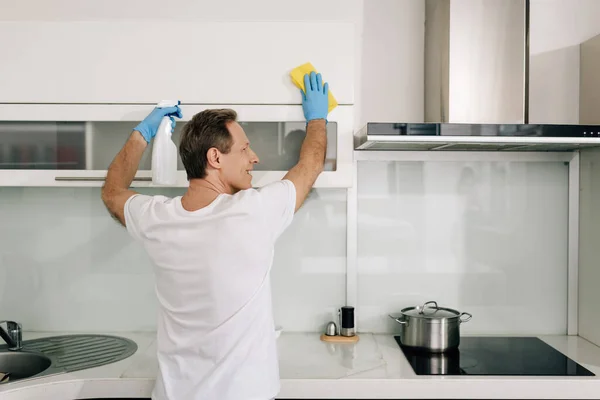 Homme heureux dans des gants en caoutchouc tenant bouteille de pulvérisation et chiffon tout en nettoyant la cuisine — Photo de stock