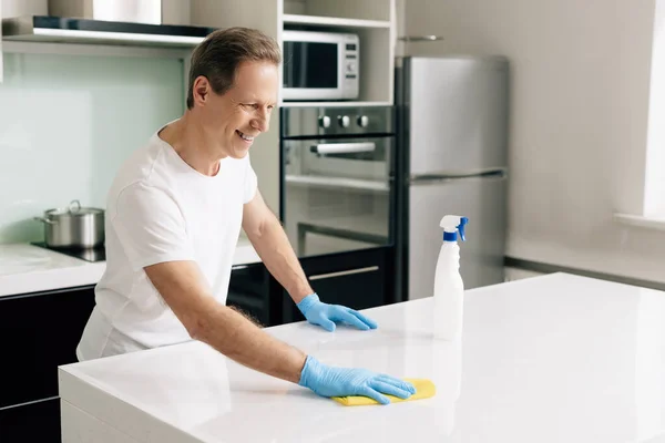 Fröhlicher Mann in Gummihandschuhen, der Lappen hält, während er Küchentisch putzt — Stockfoto