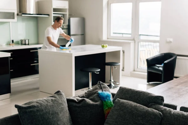 Selective focus of duster brush on sofa near man in rubber gloves cleaning kitchen table — Stock Photo