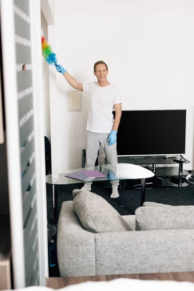 Selective focus of happy man holding duster brush while cleaning living room — Stock Photo