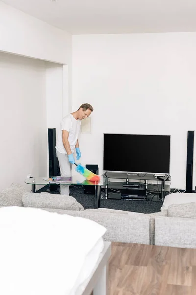 Selective focus of cheerful man holding duster brush while cleaning coffee table in living room — Stock Photo