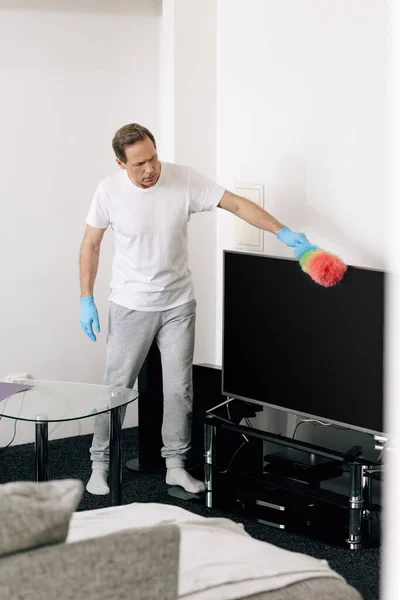 Enfoque selectivo del hombre sosteniendo cepillo de plumero mientras limpia la pantalla de televisión en blanco en la sala de estar — Stock Photo
