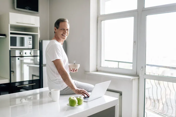 Heureux freelance tenant bol près de tasse, pommes et ordinateur portable — Photo de stock