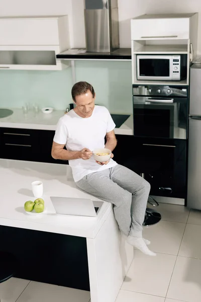 Happy freelancer holding bowl with cornflakes near cup, apples and laptop on table — Stock Photo
