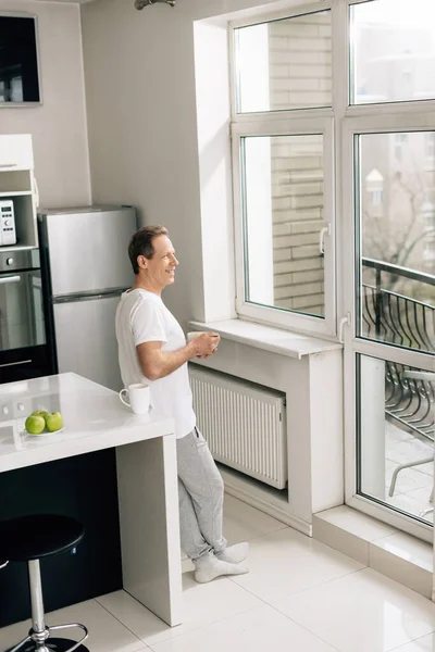Happy man holding bowl with cornflakes and looking at window near apples and cup on table — Stock Photo