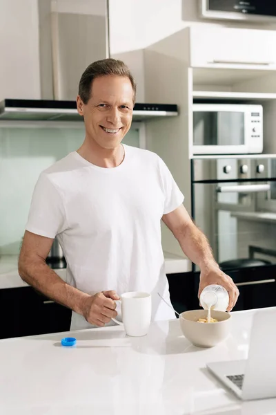 Happy freelancer pouring milk in bowl with cornflakes near laptop — Stock Photo