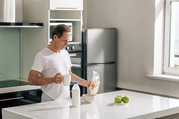 Schöner Mann gießt Cornflakes in Schüssel neben Flasche mit Milch und Äpfeln — Stockfoto