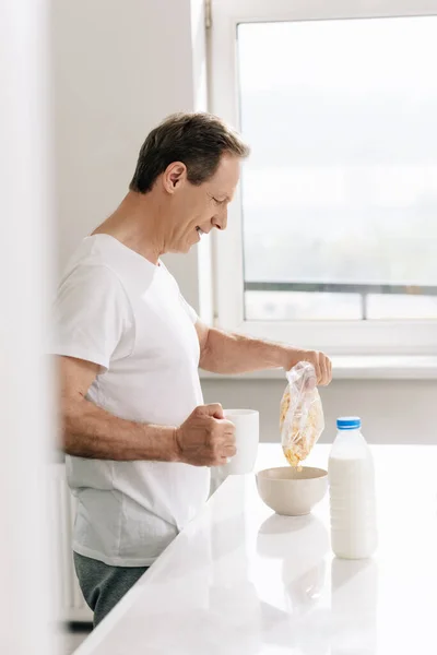 Vista lateral do homem feliz derramando flocos de milho em tigela perto de garrafa com leite — Fotografia de Stock