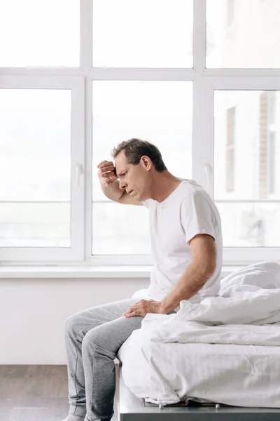 Sick man sitting on bed and touching forehead in bedroom — Stock Photo