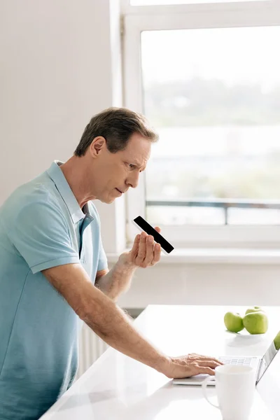 Mann hält Smartphone mit leerem Bildschirm, während er Sprachnachrichten aufzeichnet und Laptop zu Hause benutzt — Stockfoto