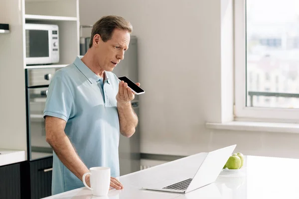 Foco seletivo do homem gravando mensagem de voz enquanto mantém o smartphone com tela preta perto do laptop — Fotografia de Stock