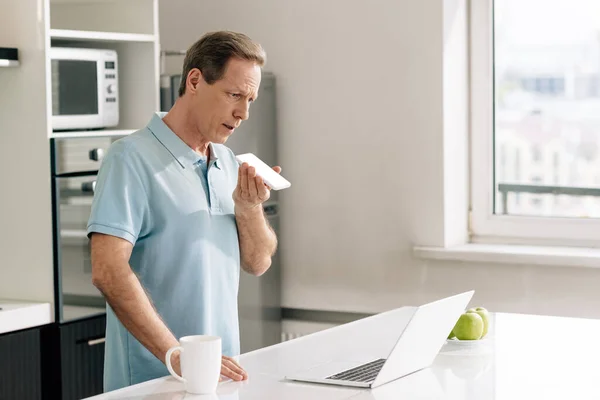 Selective focus of man recording voice message while holding smartphone with white screen near laptop — Stock Photo