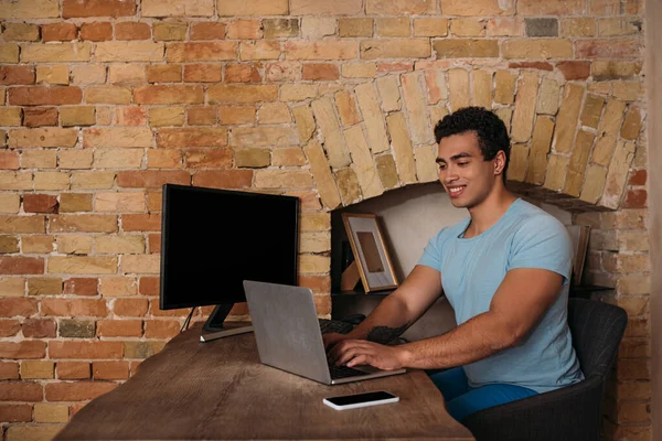 Усміхнений фрілансер змішаної раси, що працює з ноутбуком, комп'ютером і смартфоном в домашньому офісі під час самоізоляції — стокове фото