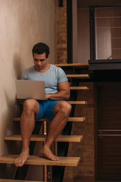 Bello uomo razza mista utilizzando il computer portatile sulle scale a casa su auto isolamento — Foto stock