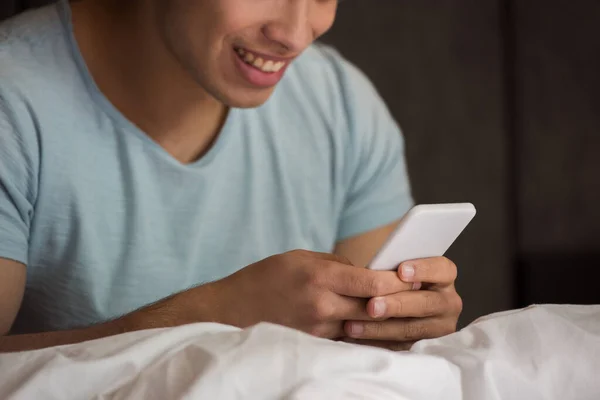 Vista recortada de hombre de raza mixta joven feliz usando smartphone en la cama en cuarentena - foto de stock