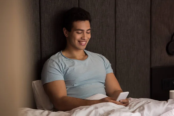 Bello sorridente uomo razza mista utilizzando smartphone a letto in quarantena — Foto stock