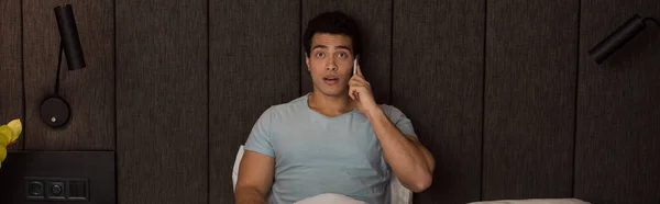 Surprised mixed race man talking on smartphone in bed on quarantine, horizontal concept — Stock Photo