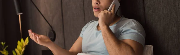 Worried mixed race man gesturing and talking on smartphone in bed on quarantine, panoramic crop — Stock Photo