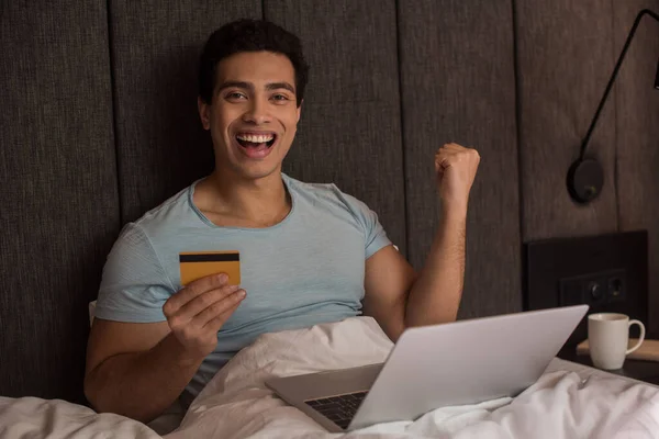 Excité mixte homme de course shopping en ligne avec carte de crédit et ordinateur portable au lit pendant l'isolement personnel — Photo de stock