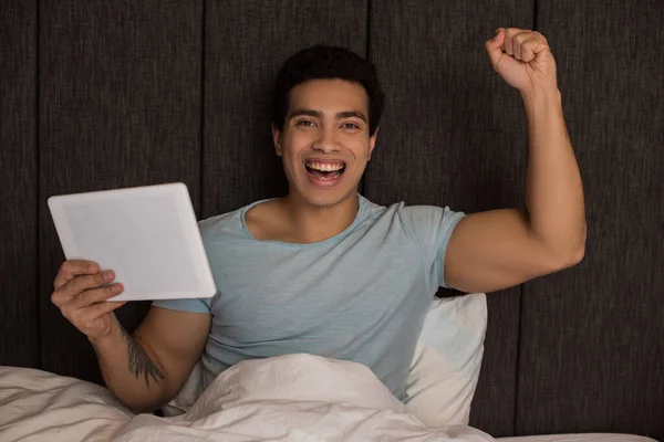 Emocionado hombre de raza mixta utilizando tableta digital en la cama durante el auto aislamiento - foto de stock